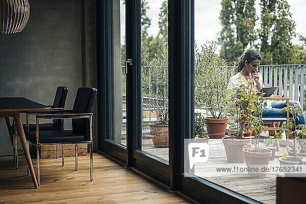 Frau sitzt mit Tablet-PC auf Balkon und sieht durch Glasfenster