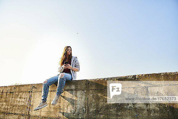 Young woman sitting on concrete wall holding smartphone