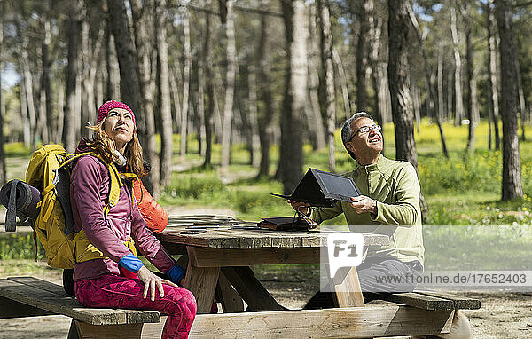 Smiling man adjusting solar panel sitting with friend at picnic table in forest