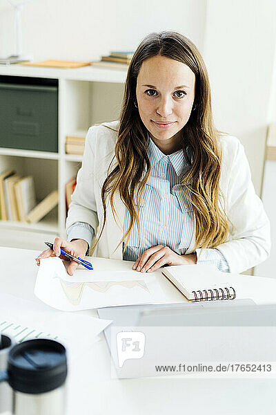 Smiling businesswoman holding pen sitting with paper at desk in office