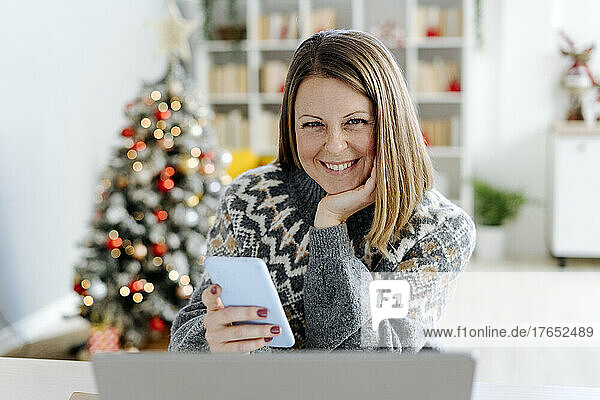 Happy woman with hand on chin holding mobile phone sitting in living room at home