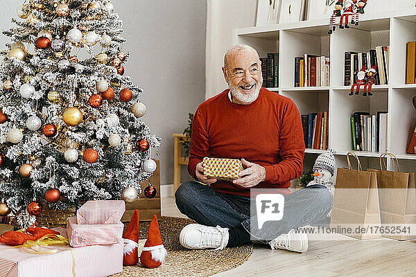 Glücklicher älterer Mann hält eine Geschenkbox in der Hand und sitzt zu Hause am Weihnachtsbaum
