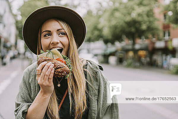 Junge Frau mit Hut isst Sandwich auf der Straße