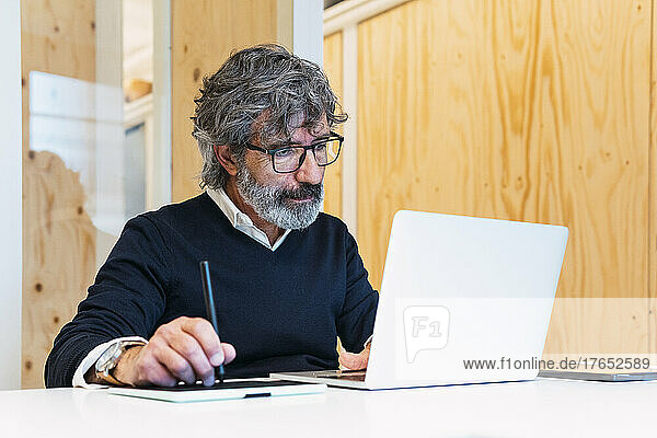 Senior freelancer using digitized pen on tablet PC looking at laptop working from home