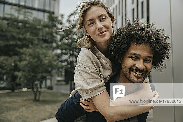 Smiling boyfriend giving piggyback ride to girlfriend in front of modern building