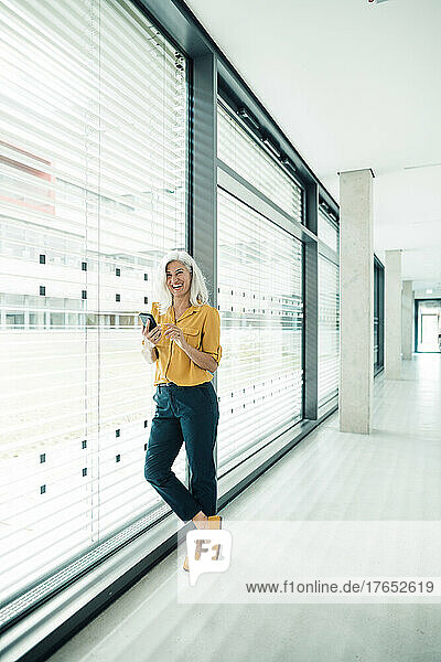 Cheerful businesswoman with smart phone leaning on glass wall in office