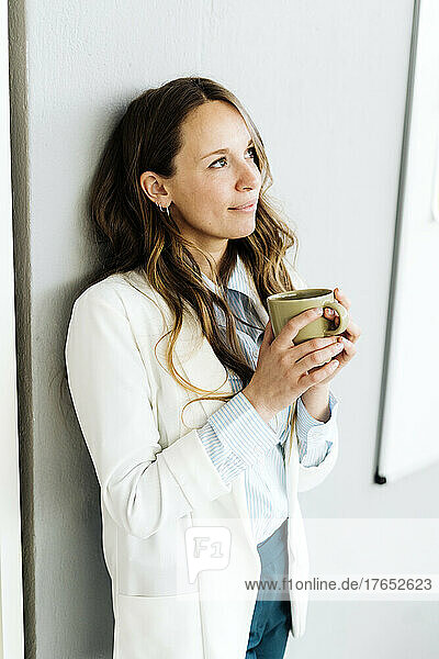 Businesswoman holding coffee cup leaning on wall at office