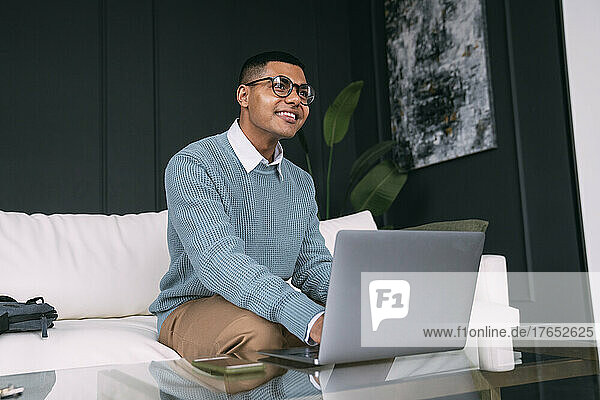 Happy freelancer wearing eyeglasses sitting with laptop on sofa at home