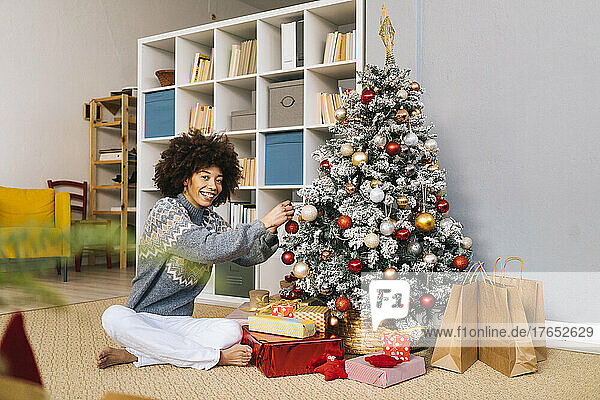 Happy woman sitting cross-legged decorating Christmas tree with bauble at home