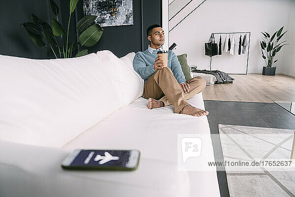 Young man with disposable coffee cup sitting on sofa in living room
