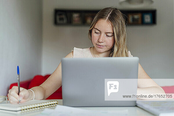 Smiling young woman writing notes sitting with laptop at home