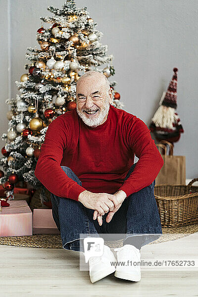 Glücklicher älterer Mann  der zu Hause vor dem Weihnachtsbaum sitzt