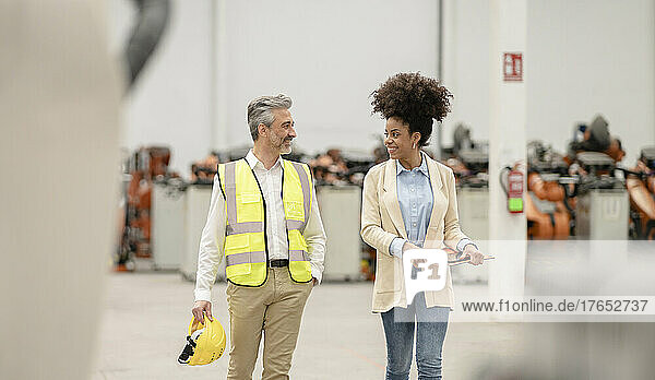 Smiling engineer holding hardhat walking with businesswoman in factory