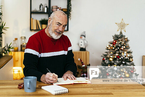 Senior man writing in diary sitting at table in living room