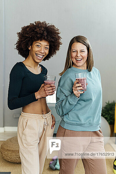 Cheerful women with glasses of smoothie standing together in living room at home