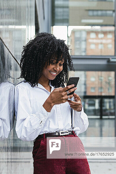 Smiling businesswoman using mobile phone leaning on wall