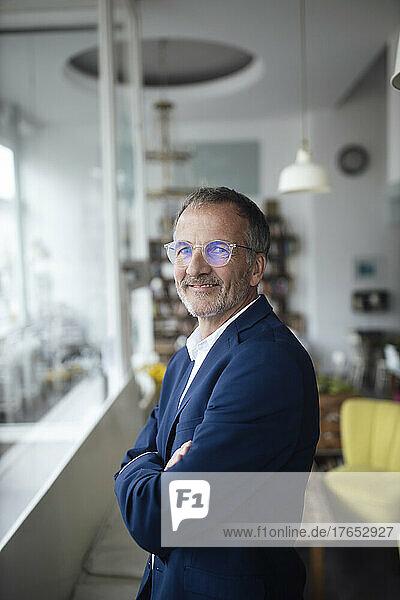 Smiling businessman wearing eyeglasses standing with arms crossed in cafe