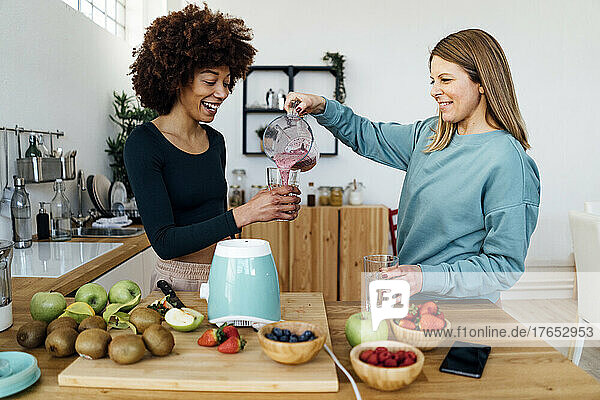 Smiling blond woman pouring smoothie to friend in kitchen at home