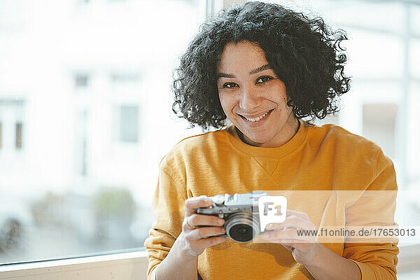 Lächelnde Frau mit Kamera sitzt zu Hause am Fenster