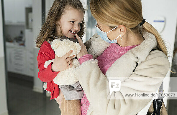 Mother carrying smiling daughter holding stuffed toy at dental clinic