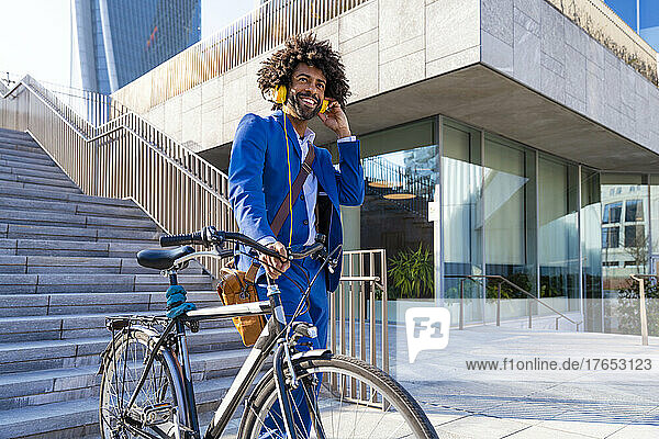 Smiling businessman with bicycle listening music through headphones on sunny day