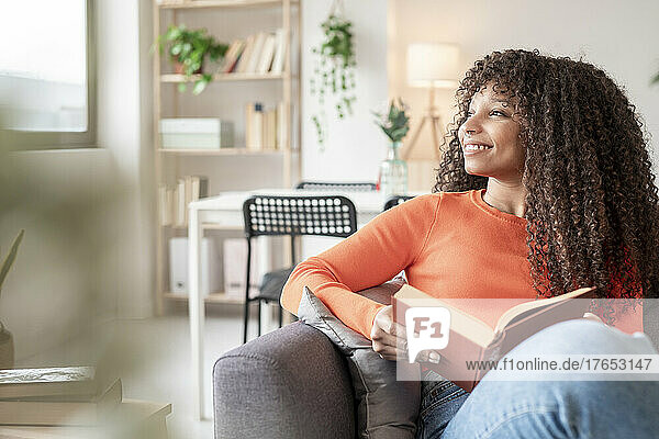 Happy young woman with book sitting on sofa in living room at home