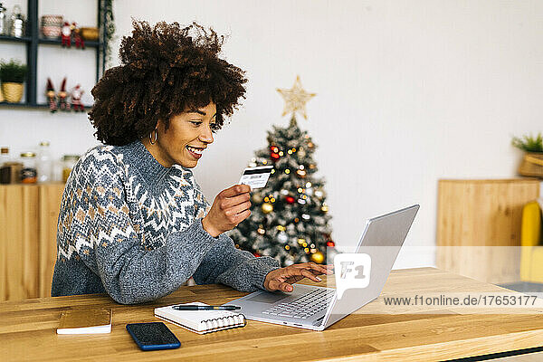 Happy young woman with credit card using laptop sitting at table in living room