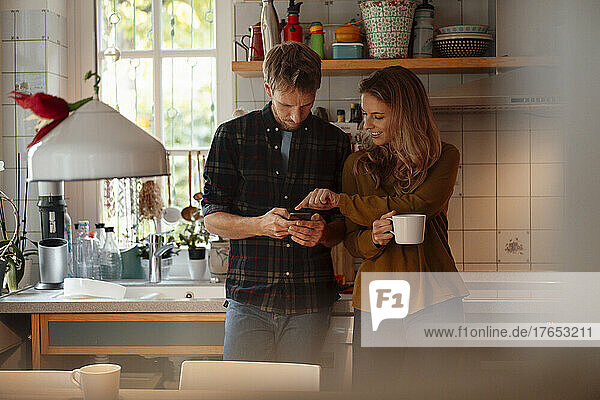 Woman holding coffee cup pointing at smart phone used by boyfriend standing in kitchen