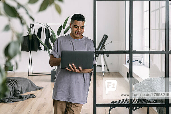 Smiling man using laptop leaning on glass wall at home