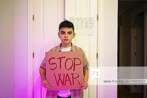 Boy with stop war placard standing in front of door at home