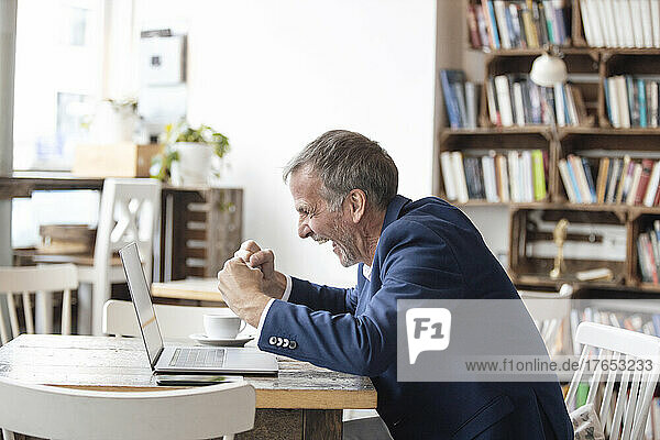 Aufgeregter Geschäftsmann sitzt mit Laptop am Tisch und feiert Erfolg im Café