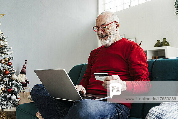 Lächelnder älterer Mann mit Kreditkarte beim Online-Einkauf sitzt zu Hause auf dem Sofa