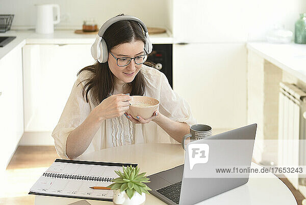 Freelancer having breakfast sitting with laptop in kitchen at home