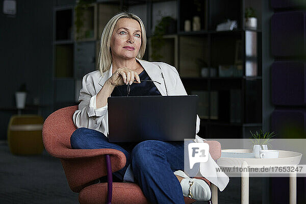 Businesswoman holding eyeglasses sitting with laptop on chair in office