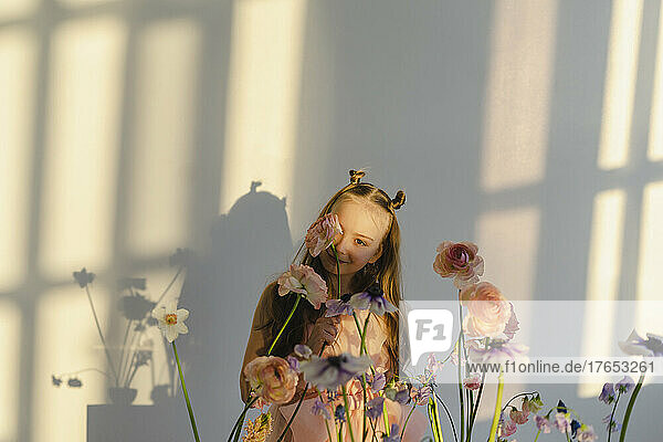 Smiling girl holding flower on face standing by white wall