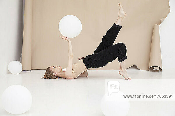 Young woman with balloon on hand exercising yoga by brown backdrop