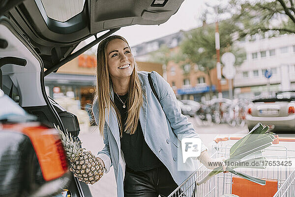 Happy young woman with shopping cart loading groceries in car trunk