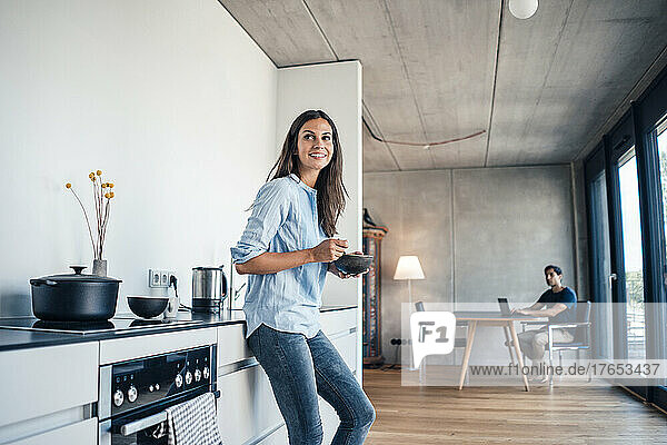 Smiling woman with bowl standing in kitchen with freelancer using laptop at home