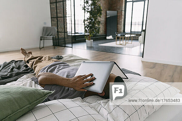 Man covering face with laptop lying on bedding at home