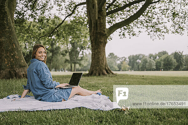 Smiling woman with laptop sitting on picnic blanket at park