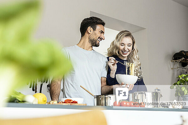 Happy man holding bowl of noodles standing with girlfriend in kitchen