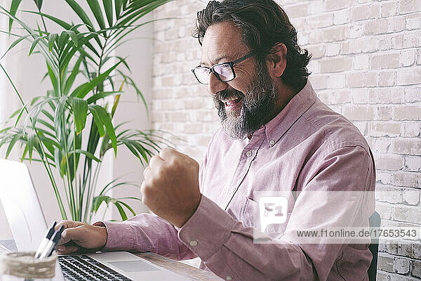 Cheerful businessman gesturing fist sitting with laptop at home