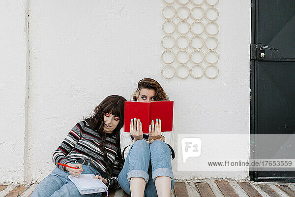Woman peeking from behind book by friend leaning head on shoulder in front of white wall