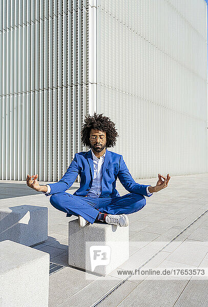 Businessman with eyes closed exercising lotus yoga pose sitting on concrete block