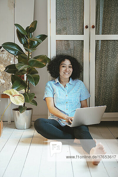 Smiling woman with laptop sitting on floorboard at home