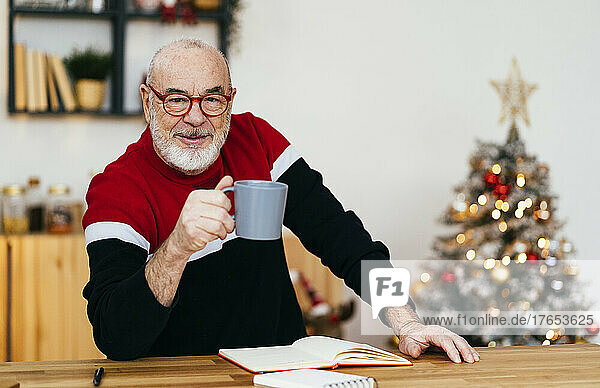 Smiling senior man wearing eyeglasses holding coffee cup sitting at table