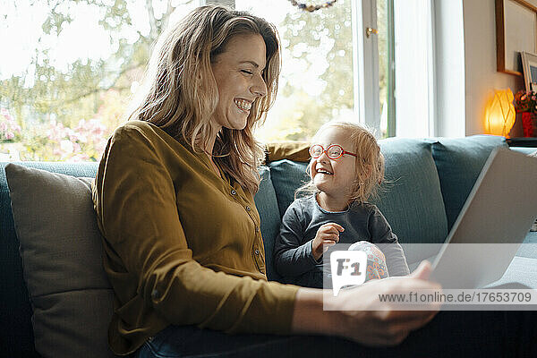 Cheerful blond woman holding tablet PC sitting by daughter on sofa at home