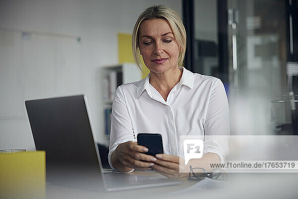 Smiling businesswoman using smart phone and laptop at desk in office