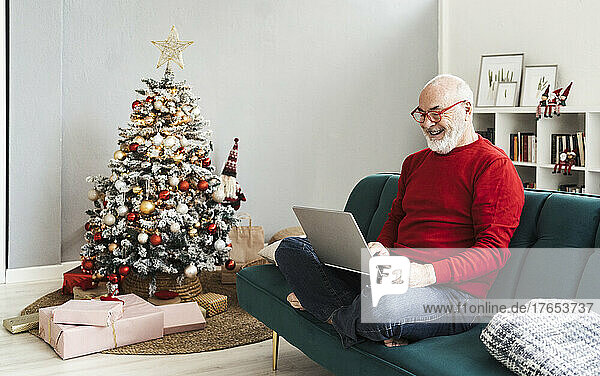 Lächelnder älterer Mann mit Laptop sitzt zu Hause auf dem Sofa im Wohnzimmer