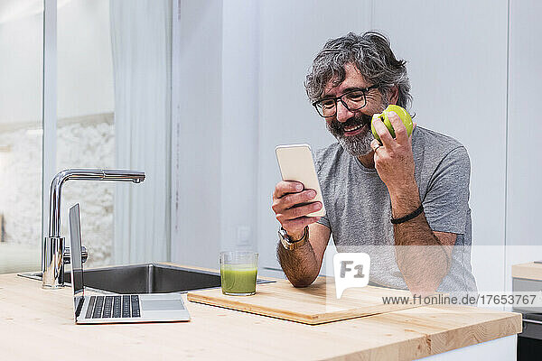 Happy senior man with apple using smart phone at kitchen island